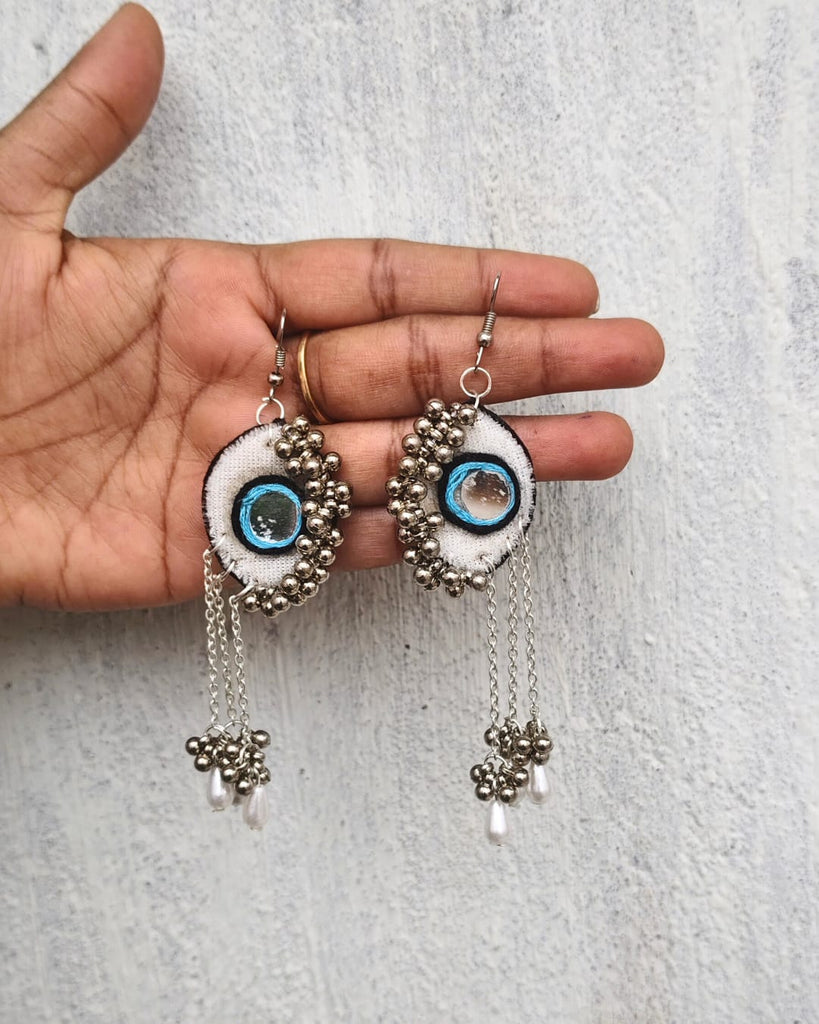 Pair of decorative earrings with eye design held by a hand on a light gray background