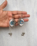Pair of decorative earrings with eye design held by a hand on a light gray background